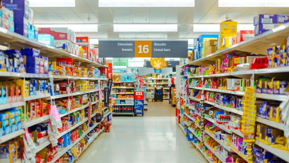 Corredor iluminado de um supermercado (corredor 16: Chocolates, Biscoitos e Doces) com prateleiras cheias de produtos coloridos.