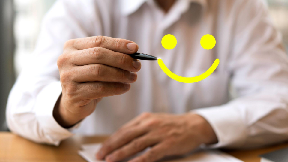 Um homem vestindo camisa social branca está sentado em uma mesa com papéis. Ele segura uma caneta preta e parece "desenhar" ou "apontar" no ar para um emoji amarelo brilhante de um rostinho sorridente (smile).