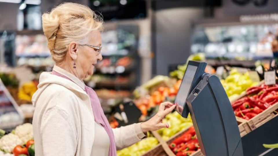 A imagem mostra uma mulher idosa em um supermercado, no setor de hortifrúti, utilizando uma balança digital com tela sensível ao toque, com inteligência artificial.