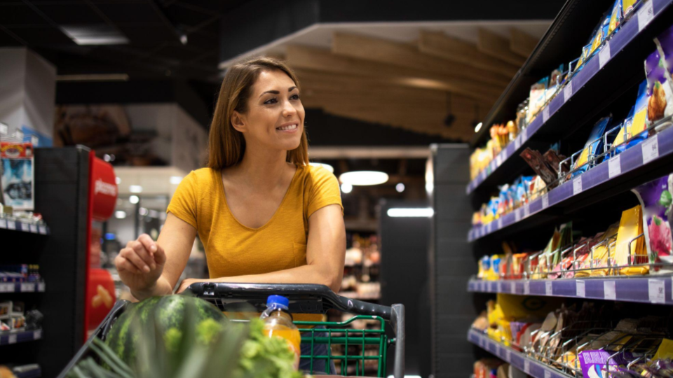 Uma mulher de cabelos castanhos claros está no corredor de um supermercado, sorrindo suavemente enquanto olha para os produtos nas prateleiras à sua direita.
