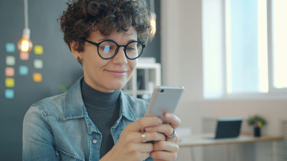 Uma mulher de cabelo curto e cacheado sorri suavemente enquanto segura e olha para a tela de um smartphone prateado fazendo compras.