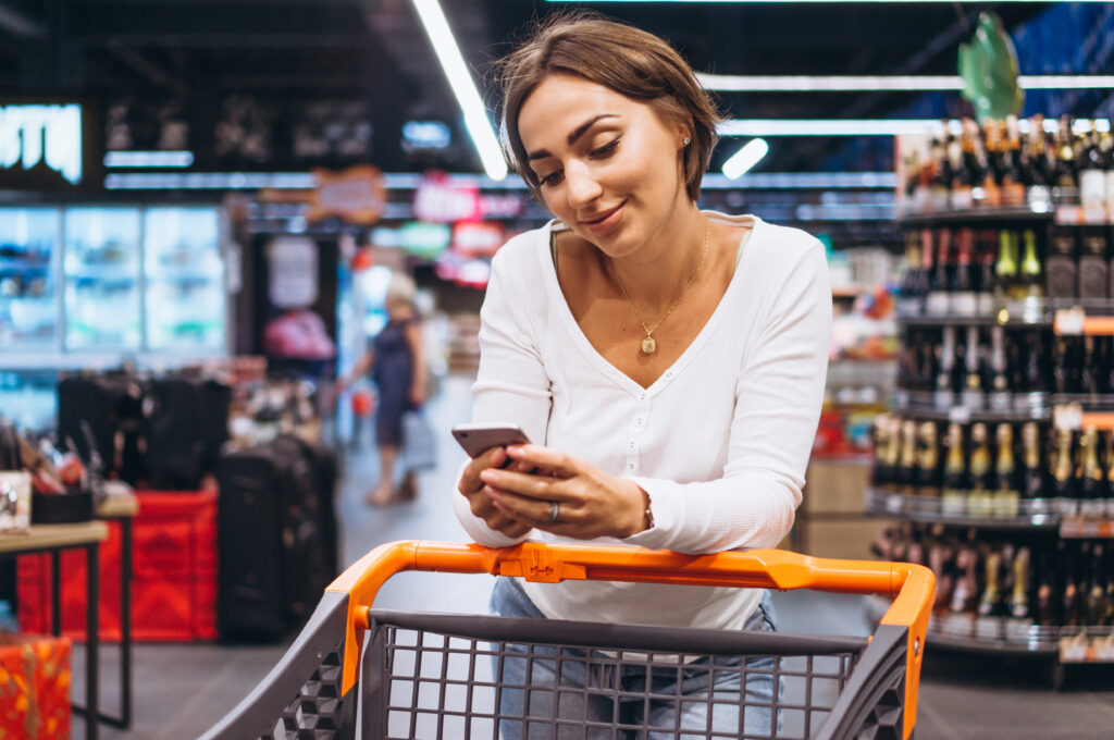A imagem mostra uma mulher de cabelo curto castanho no corredor de um supermercado, debruçada sobre o apoio de um carrinho de compras cinza com alça laranja.
