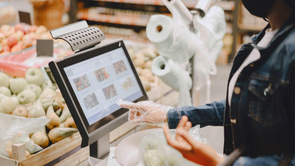 A imagem mostra uma pessoa no setor de hortifrúti de um supermercado a utilizar uma balança digital com ecrã tátil.